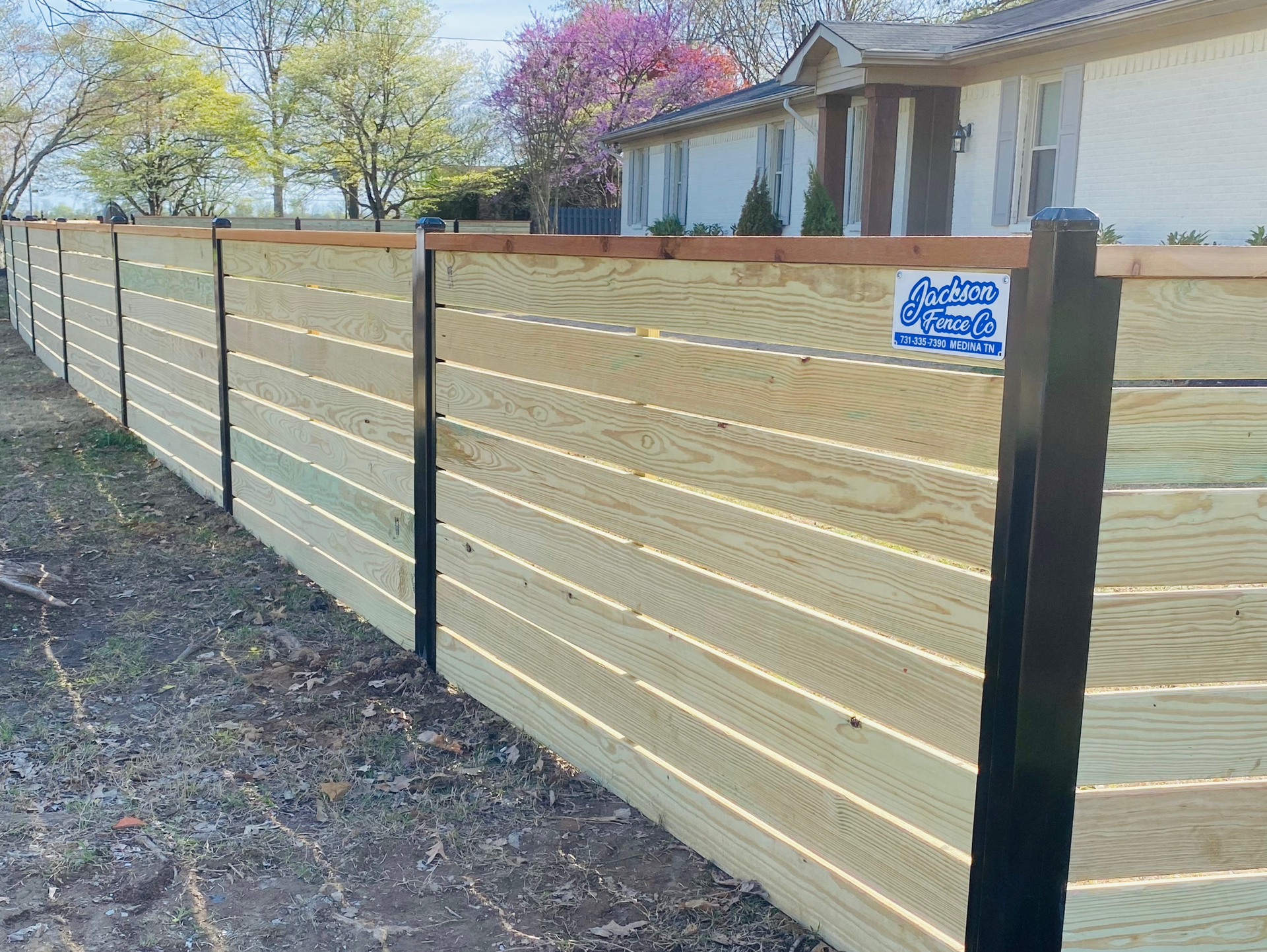 New wooden privacy fence along a yard beside a house, with black posts and a blue sign.