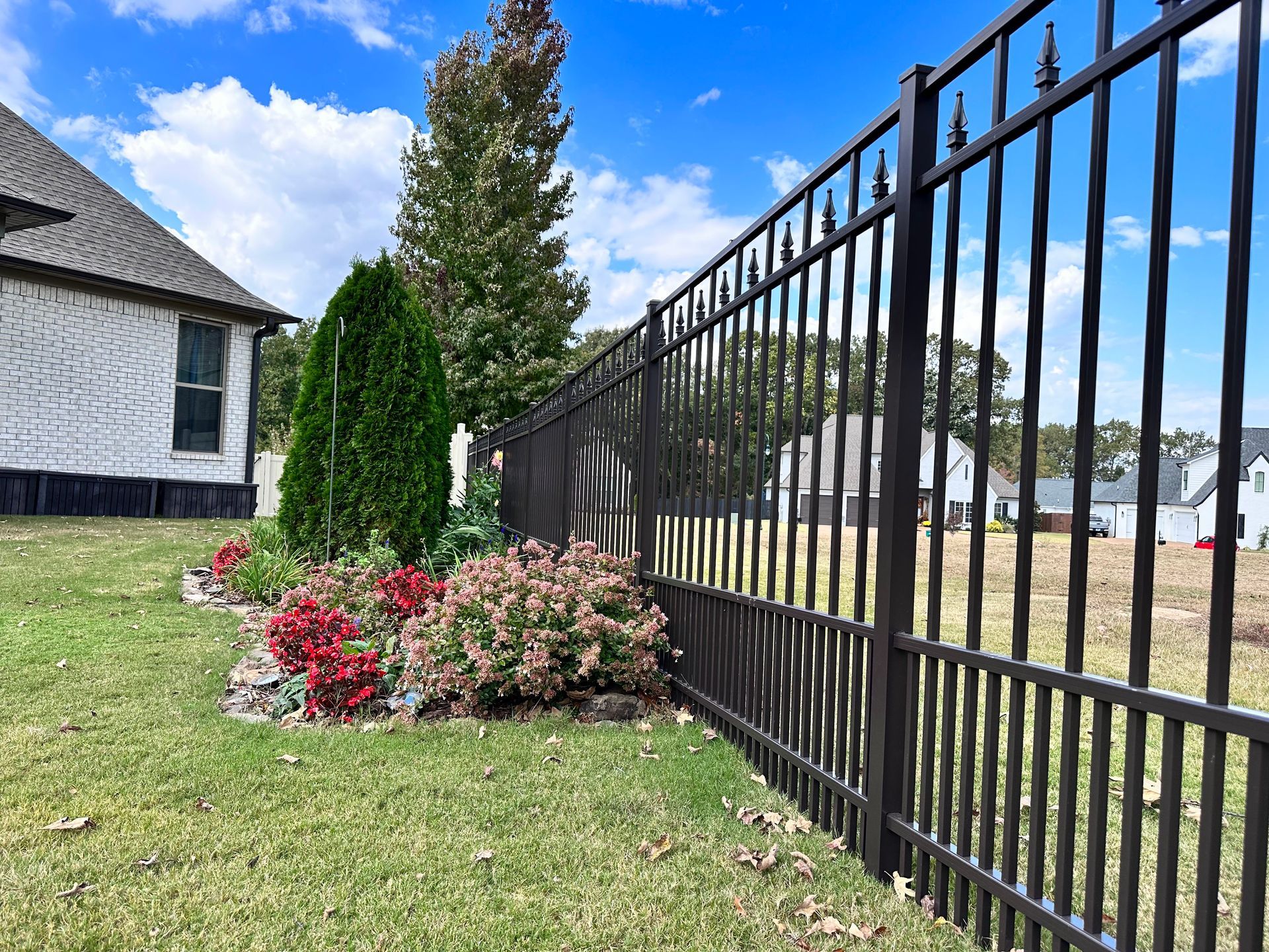 Black metal fence beside a house, with shrubs and flowers along a grassy yard under a blue sky.