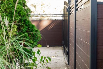 Narrow outdoor walkway with brown fence, concrete path, and green plants beside a house