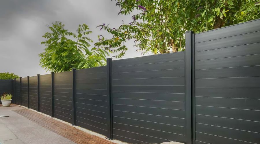 A tall, modern horizontal black privacy fence lines a paved walkway with green trees in the background.