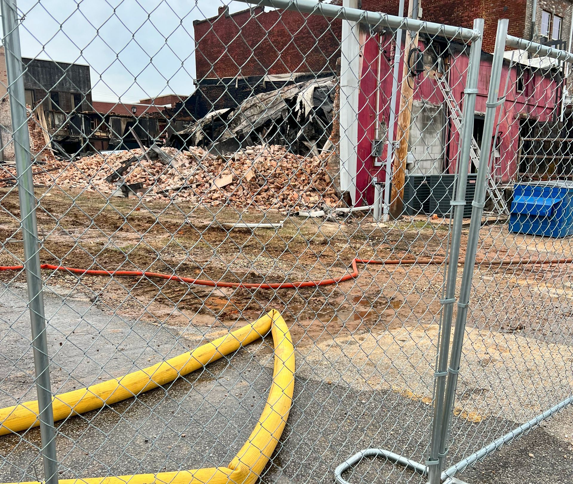 Collapsed building behind chain-link fence with rubble, orange safety mesh, and yellow hose in foreground