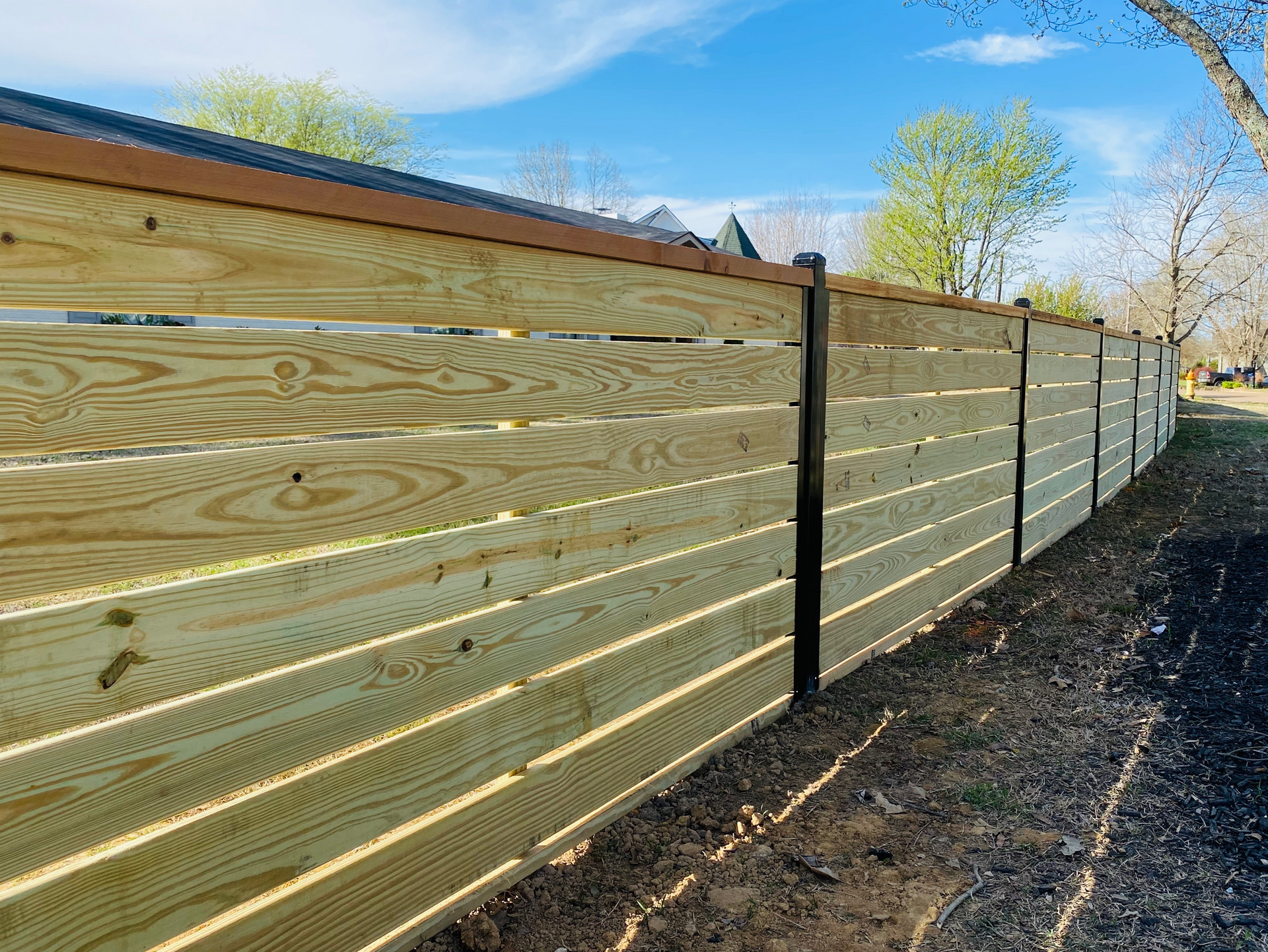 Wooden privacy fence with black posts along a gravel path under a blue sky