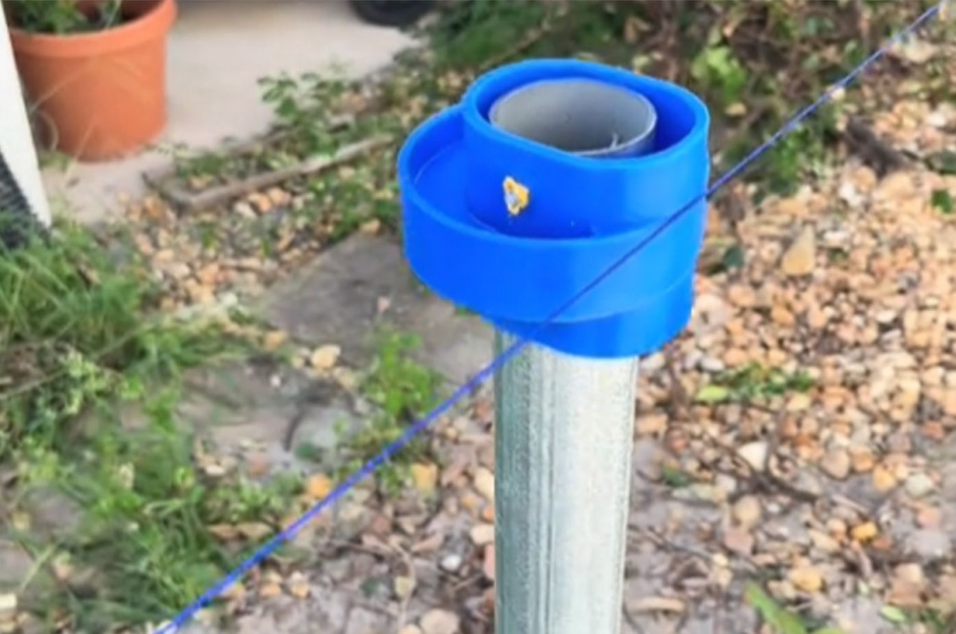 Blue plastic wrap around a metal post in a gravel yard, with a thin cord attached.