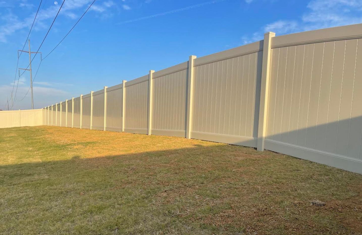 Long beige privacy fence along a grassy yard under a blue sky at sunset.