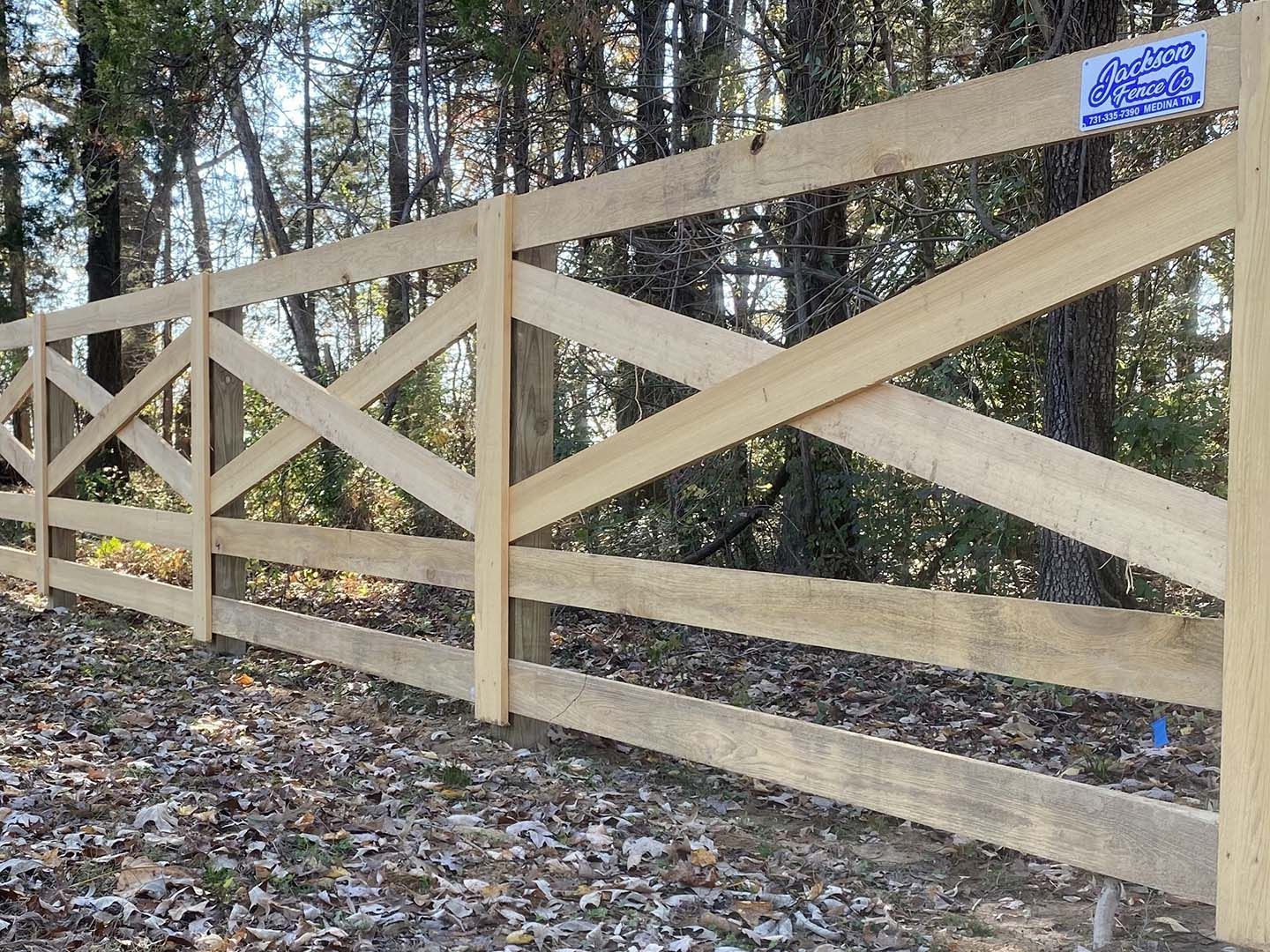 Wooden farm gate with diagonal braces along a wooded trail, with a blue sign attached near the top.