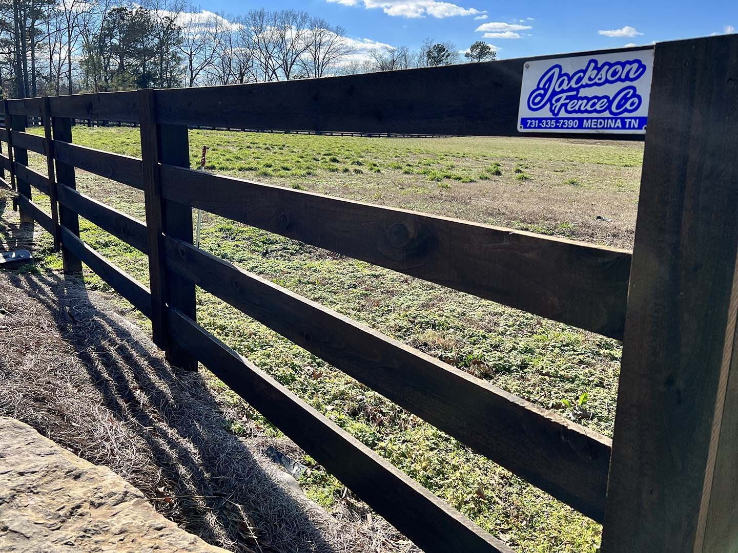 Black wooden fence beside a grassy field with a blue Jackson Fence Co. sign under a sunny sky