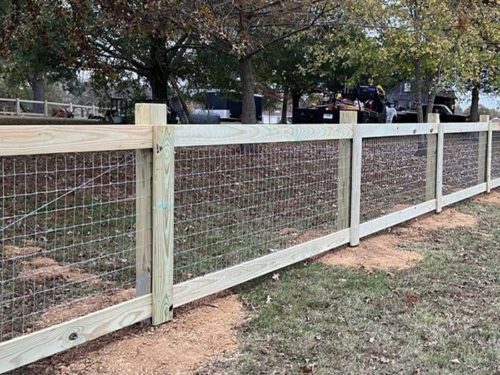 Wooden wire-mesh fence along a grassy yard with trees and parked cars in the background