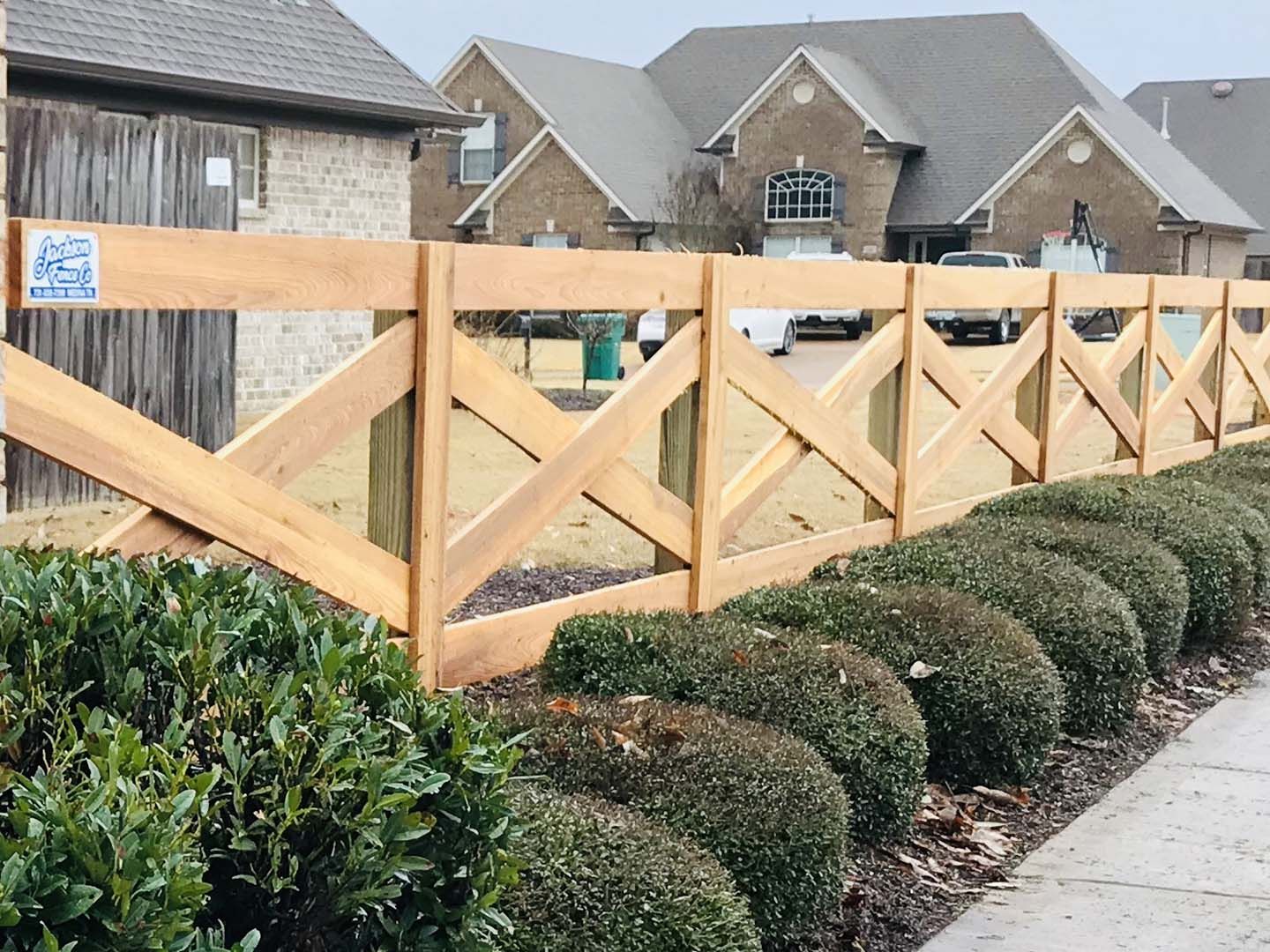 Wooden picket fence beside trimmed shrubs along a suburban sidewalk, with houses in the background