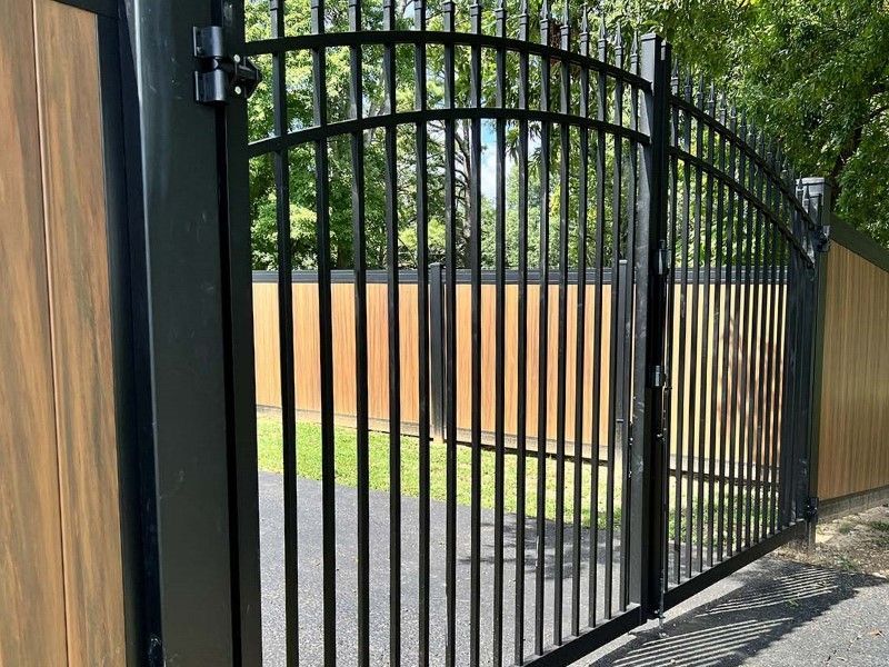 Black metal gate beside a wooden fence, opening onto a paved driveway with trees beyond