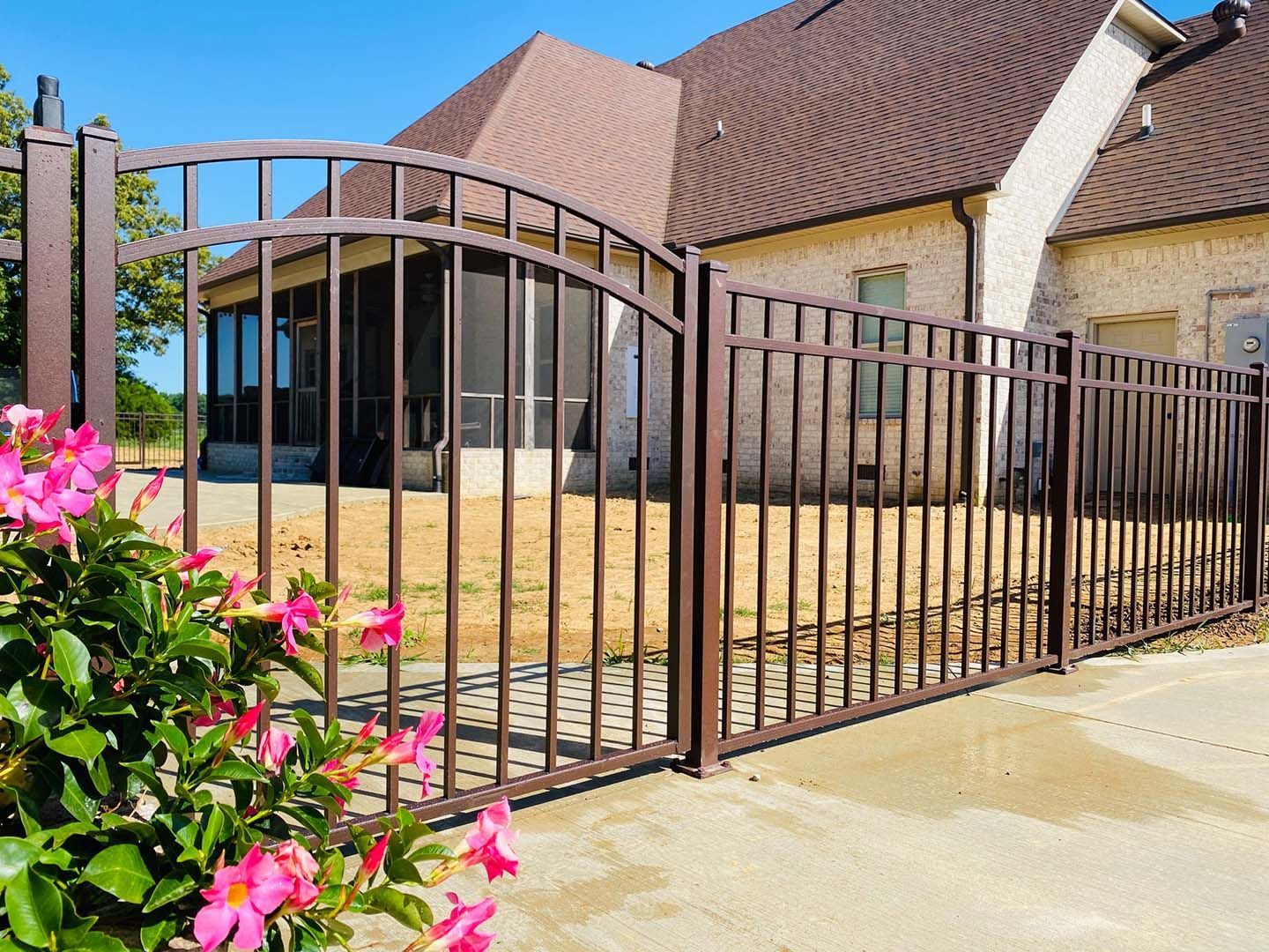 Brown metal driveway gate beside a brick house, with pink flowers in the foreground