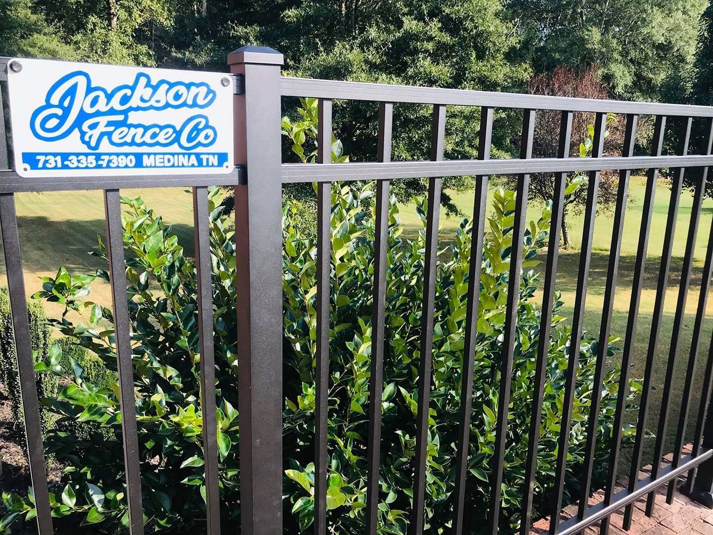 Jackson Fence Co. sign on a black metal fence by bushes and trees outdoors