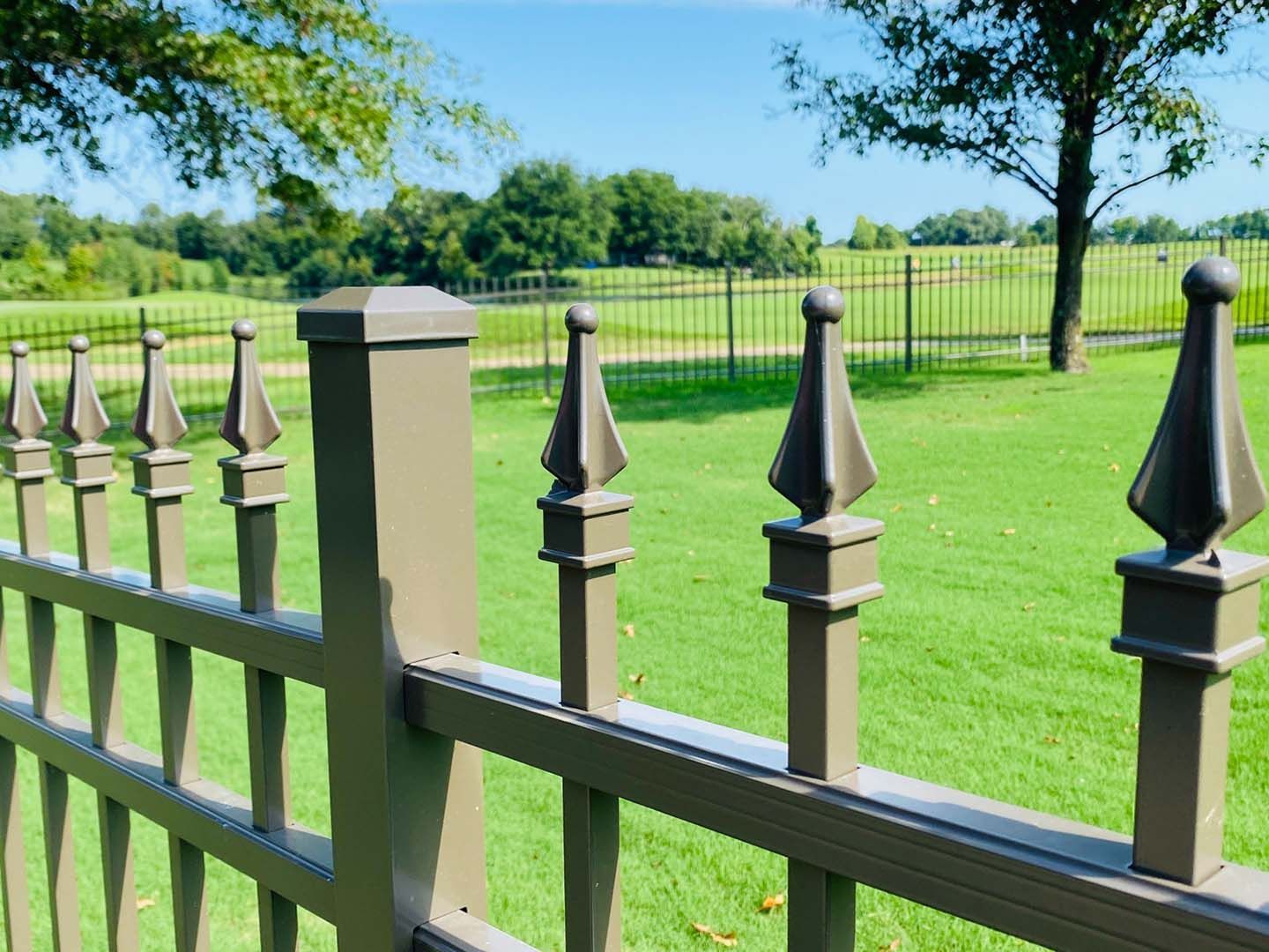 Black metal fence with pointed posts in front of a green field and trees under a bright sky