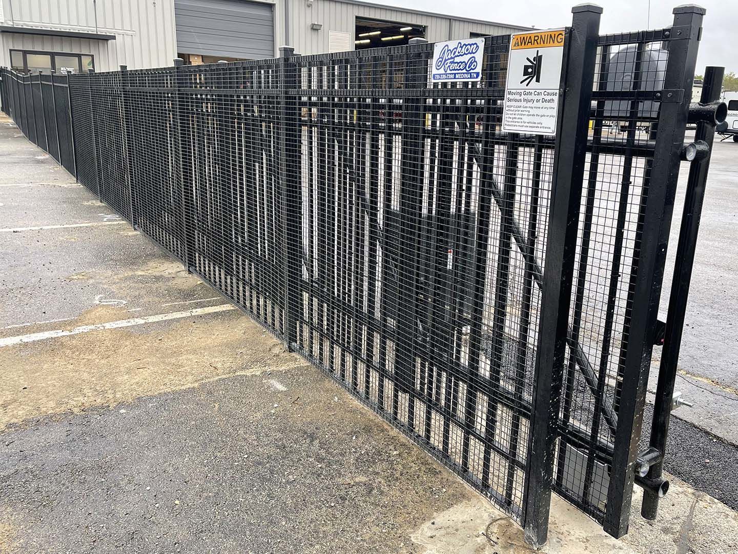 Black metal security fence and gate along a concrete walkway beside an industrial building.