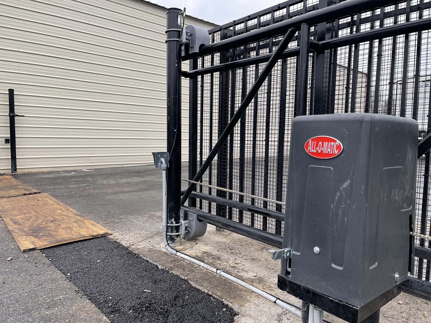 Gray gate beside building with a metal security box and a plywood ramp over uneven pavement.