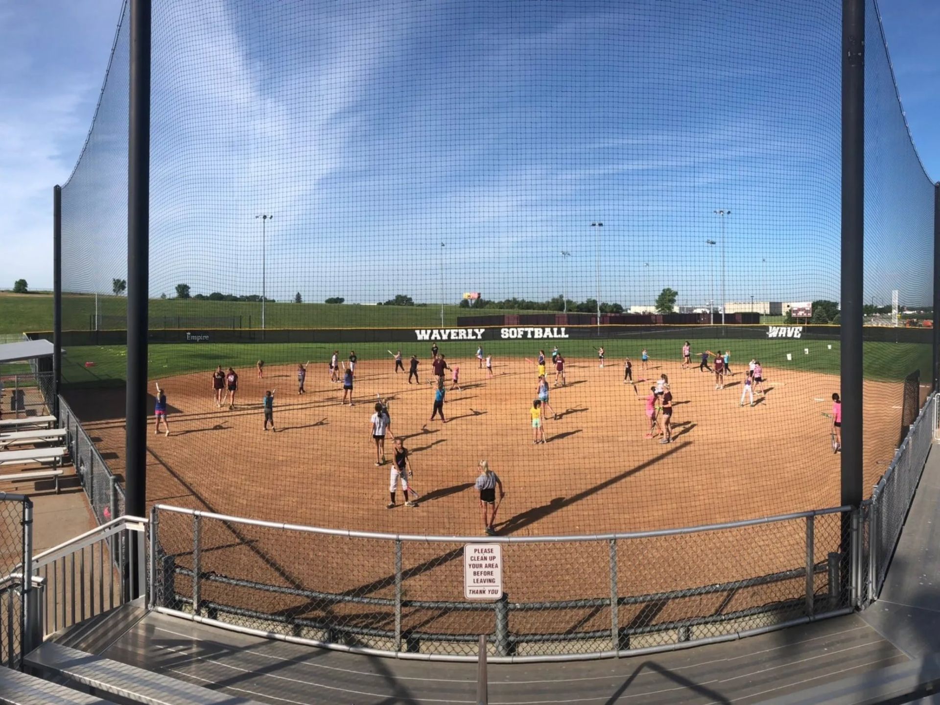 Softball game on a dirt field behind a protective net under a bright blue sky.