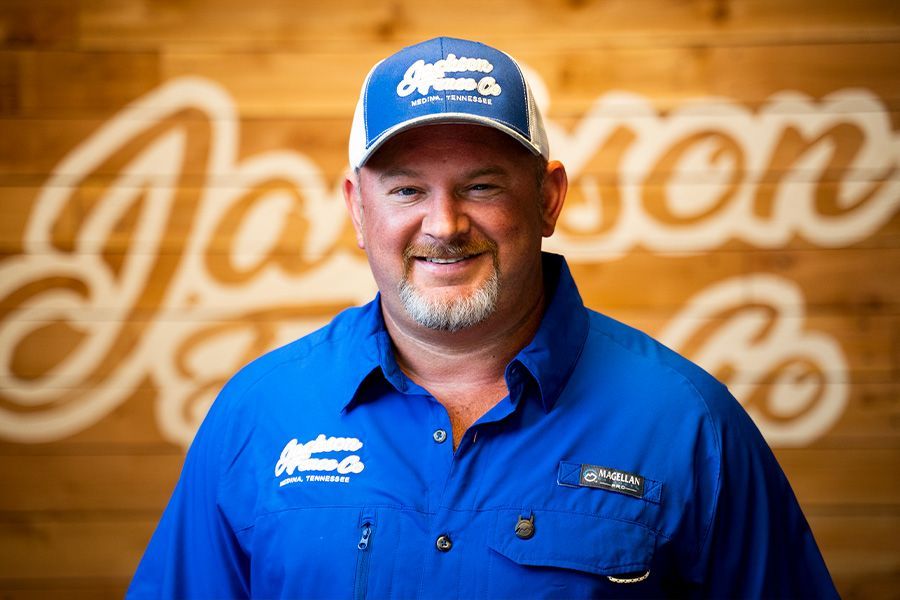 Smiling man in blue work shirt and cap standing before a wood wall with cursive signage