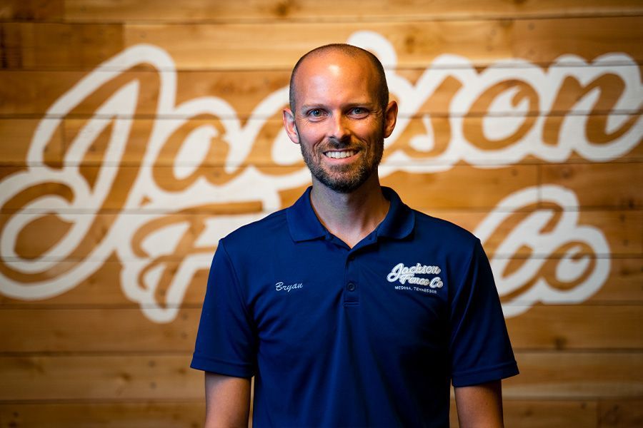Smiling bald man in a navy polo standing before a wooden wall with a white script logo