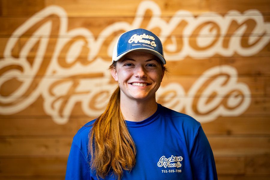 Smiling person in blue shirt and cap standing before a wooden wall with “Jackson Farm” sign