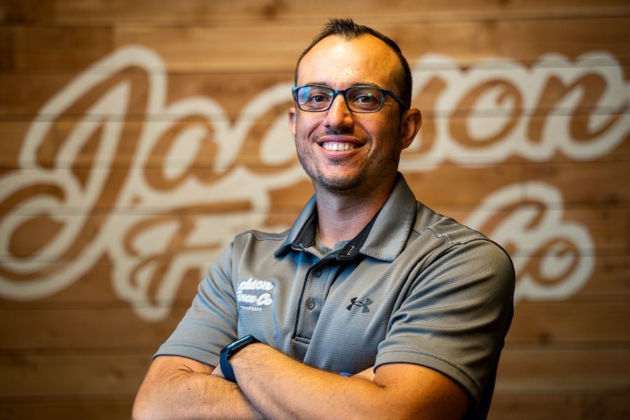 Smiling man in glasses and gray polo standing with arms crossed in front of a wooden wall logo