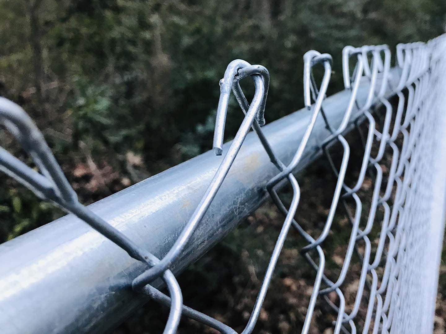 Close-up of a bent chain-link fence attached to a metal railing outdoors, with trees in the background.