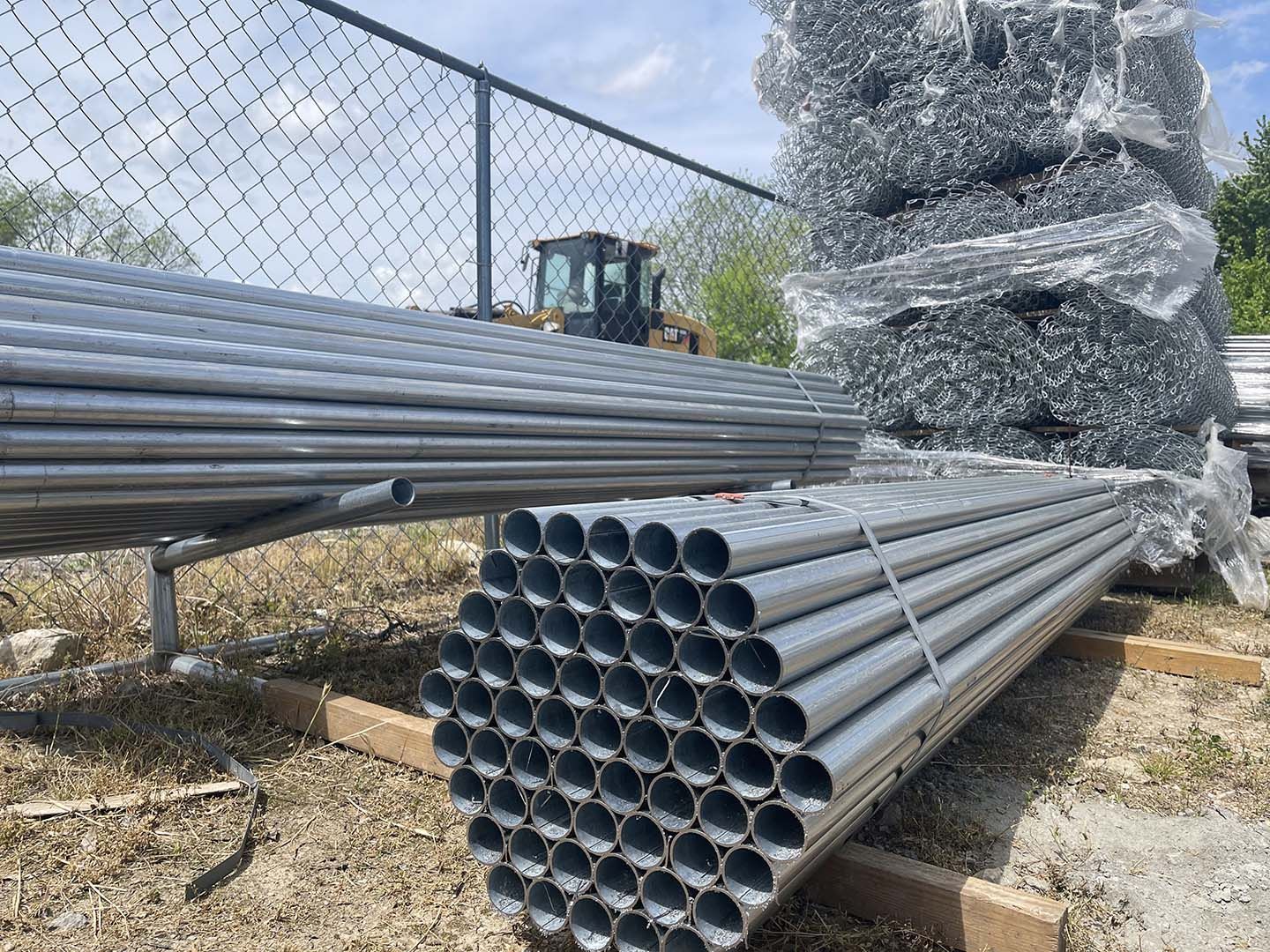 Stacked gray metal pipes and bundled wire mesh outdoors by a fence, with a forklift in the background