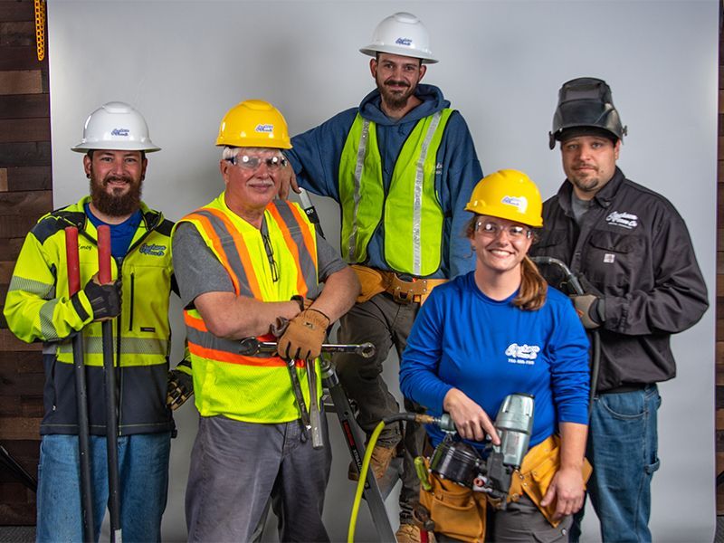 Five construction workers in hard hats and safety vests pose with tools at a job site.