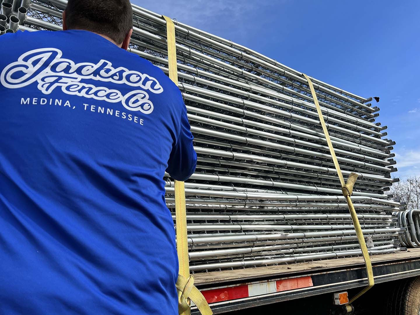 Worker in blue Jackson Fence shirt unloading stacked metal fence panels from a flatbed truck
