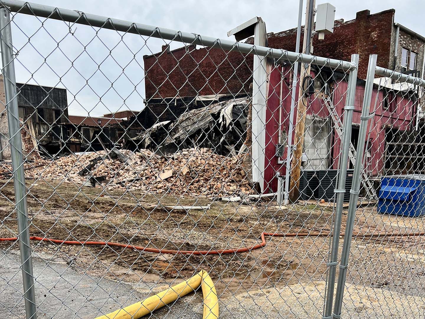 Fenced demolition site with rubble piles, damaged brick buildings, and a yellow hose on wet pavement.