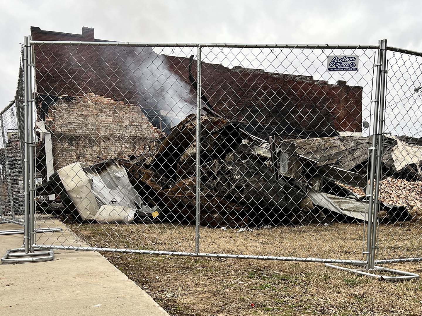 Burned-out debris behind a chain-link fence, with smoke rising from the rubble