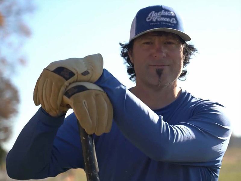 Man in a blue shirt and cap holding a golf club, wearing tan gloves outdoors