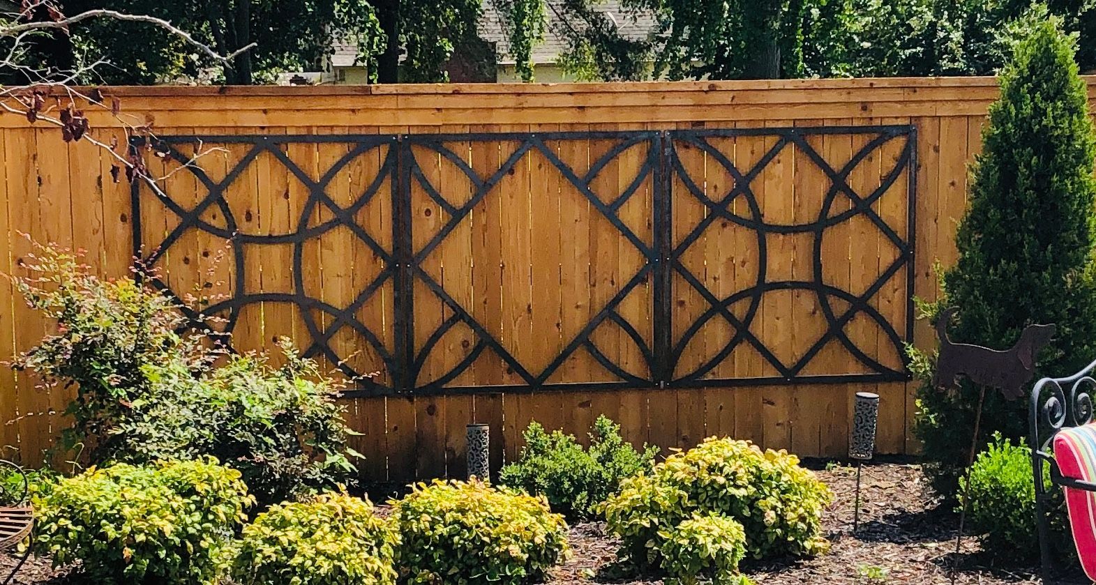Wooden garden fence with dark lattice and diamond pattern, bordered by shrubs and trees in sunlight