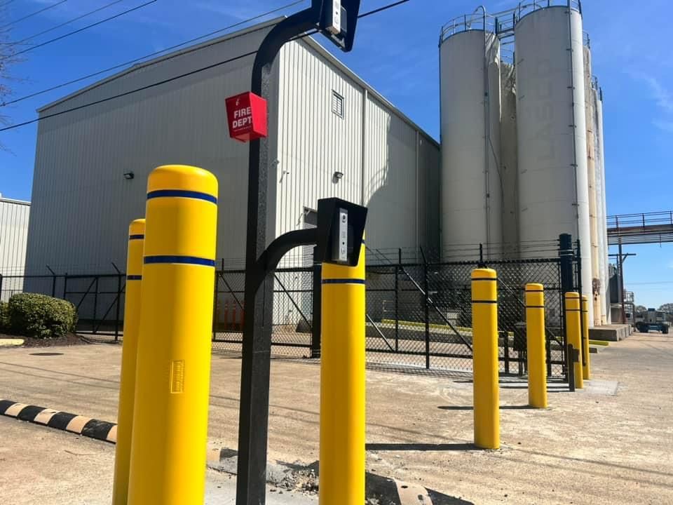 Industrial facility with yellow safety bollards beside large metal silos and a warehouse under a blue sky