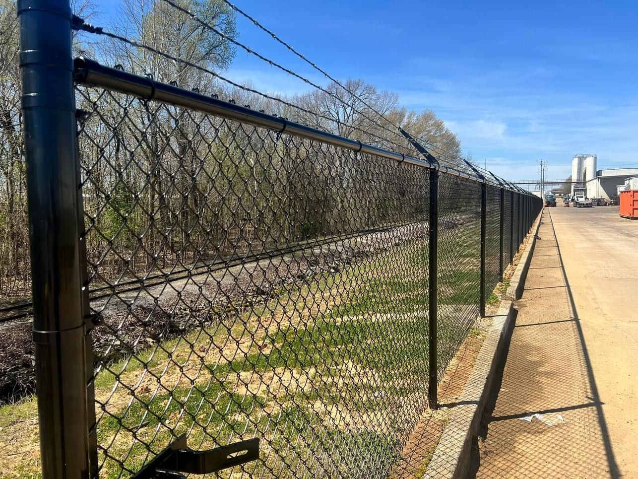 Black chain-link fence along a gravel path beside a road under a clear blue sky