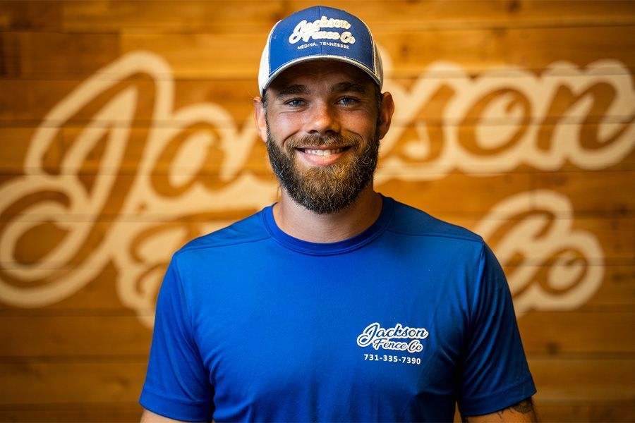 Smiling man in a blue cap and shirt stands before a wooden wall with a large script logo.