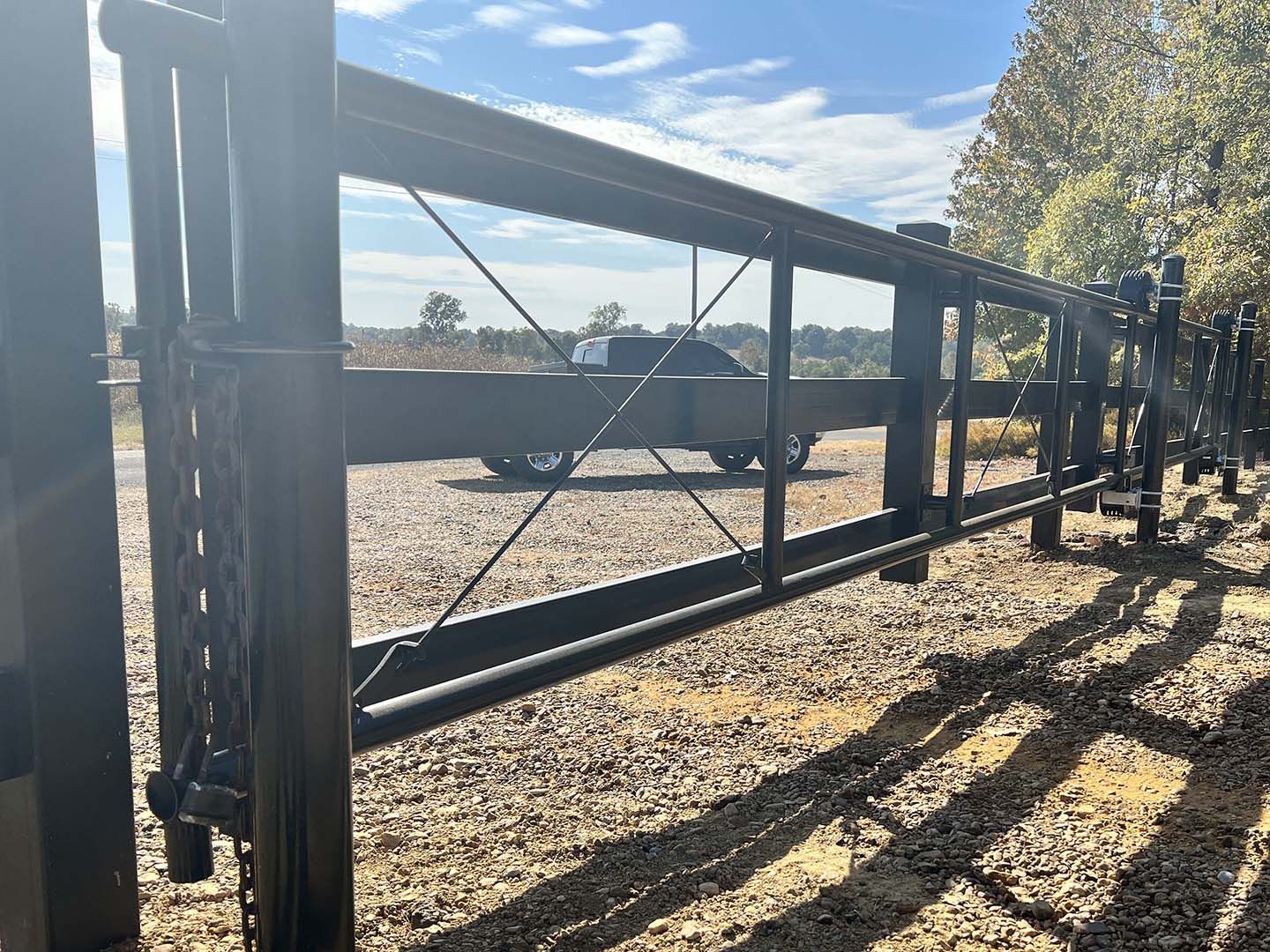 Black metal farm gate with diagonal braces along a gravel driveway in bright sunlight.