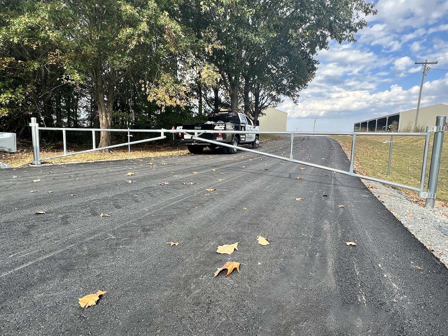 Closed gravel road with a white barrier gate, trees on the left, and a chain-link fence on the right.