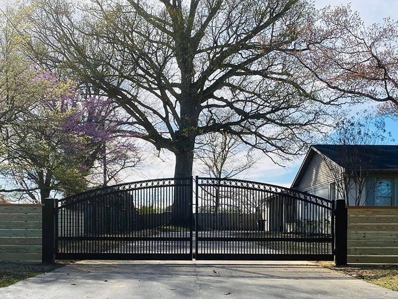 Black metal gate before a large tree and house, with blooming trees in the background