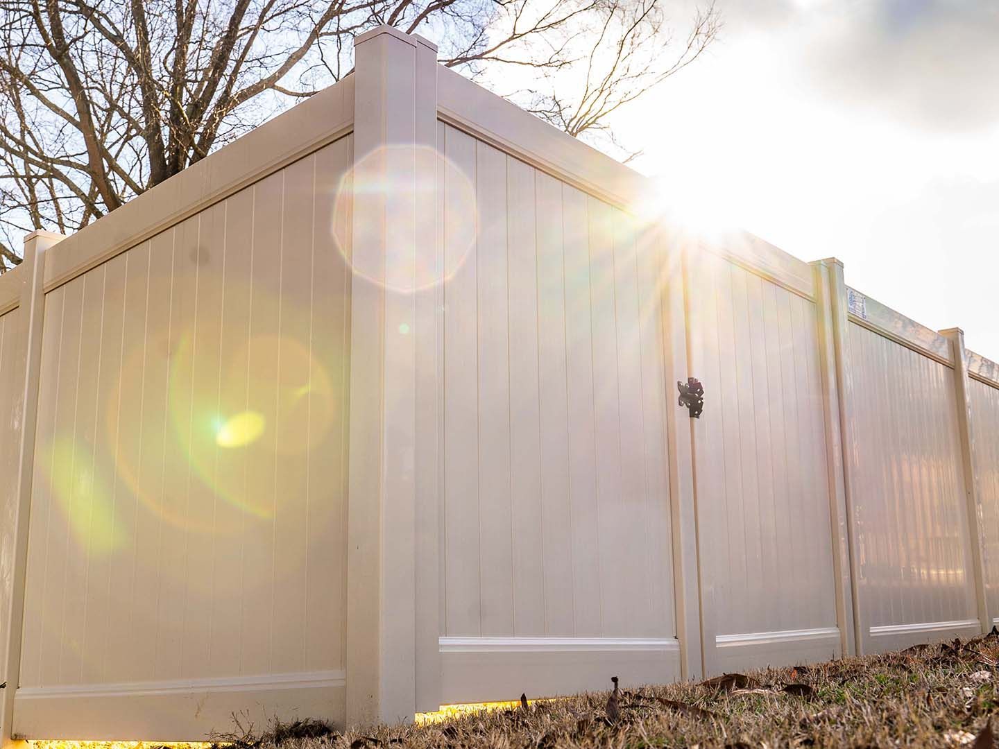 White vinyl privacy fence in bright sunlight with lens flare and bare trees behind it