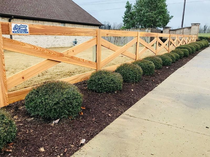 Wooden fence with diagonal braces beside trimmed shrubs along a concrete sidewalk