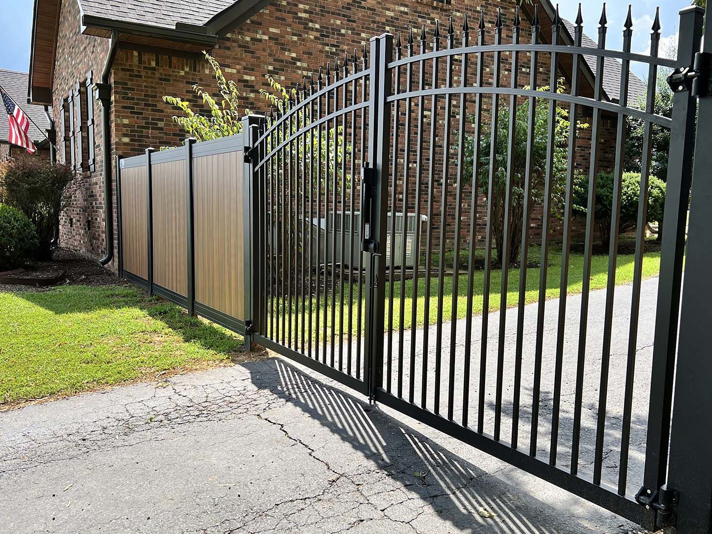 Black metal driveway gate beside a brick house and fence, opening onto a paved driveway and lawn.