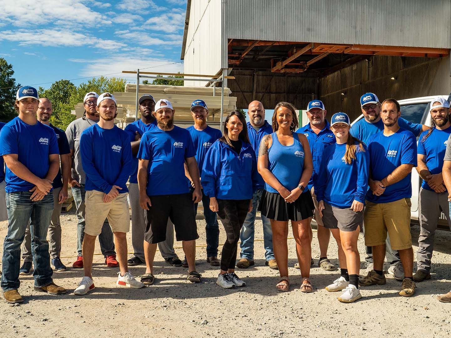 Group of volunteers in blue shirts standing outdoors in front of a warehouse-like building.