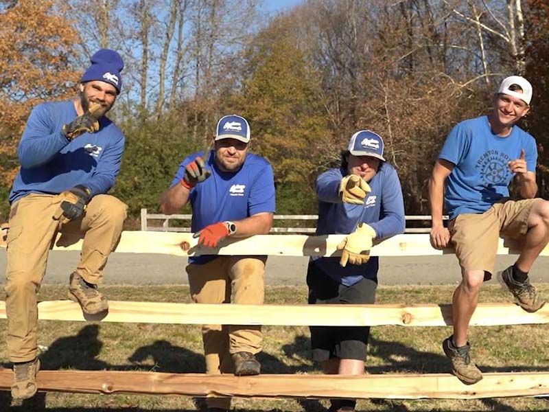 Four people in blue shirts and caps sit on a wooden fence outdoors, smiling in a sunny field.