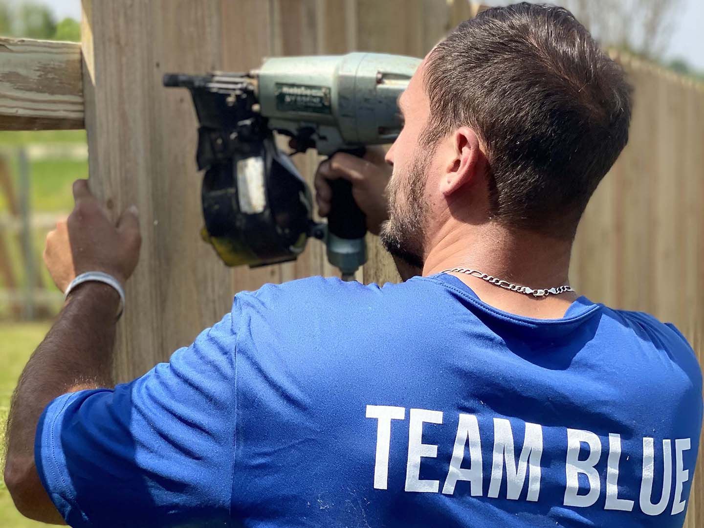 Person in blue “TEAM BLUE” shirt aiming a paintball marker at a wooden barrier.
