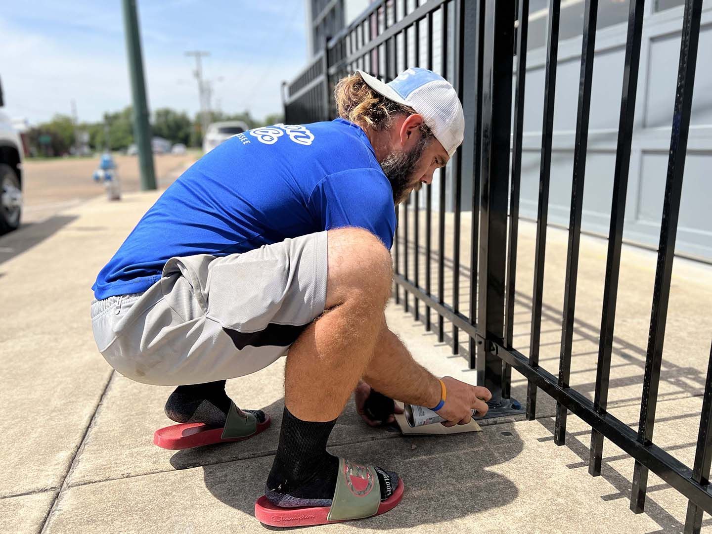 Worker crouches beside a black metal fence, tightening a bolt on a sidewalk.