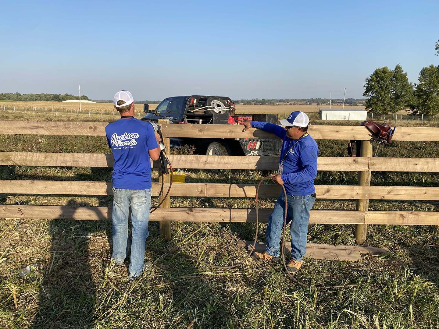 Two workers in blue shirts fixing a wooden fence in a field, with a pickup truck nearby.