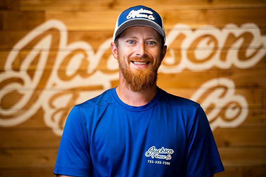 Smiling man in blue shirt and cap standing before a wooden wall with a blurred “Jayson” logo.