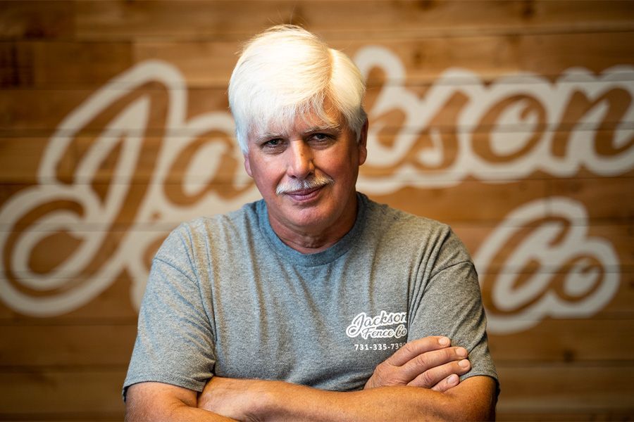 Man with white hair and crossed arms in a gray shirt, standing before a wood wall with a large script logo.