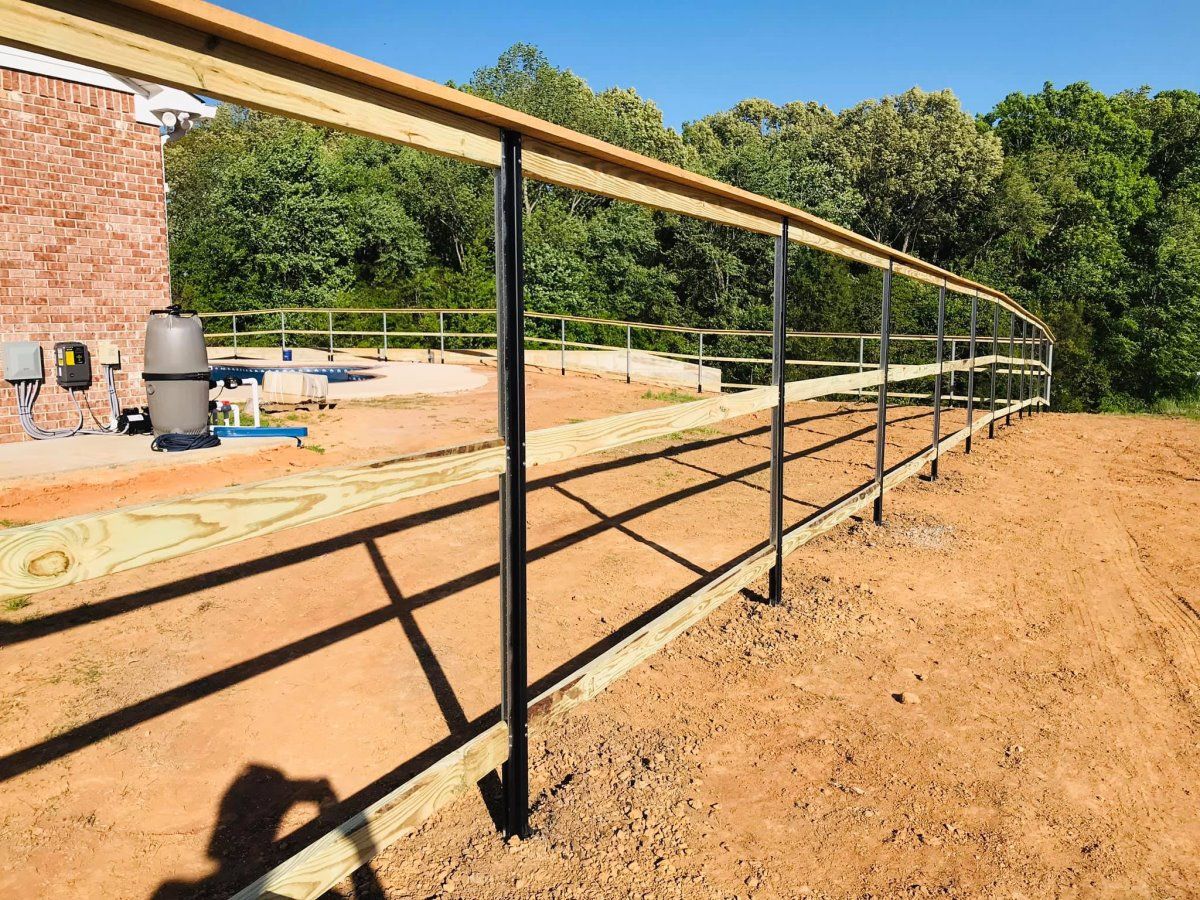 Long metal railing beside a dirt walkway under construction near a brick building and trees