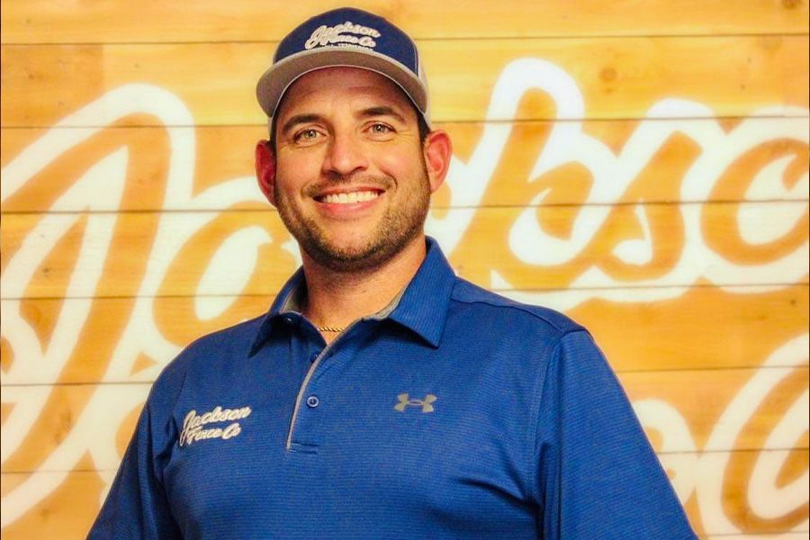 Smiling man in a blue polo and cap standing before a wood wall with a large white script logo