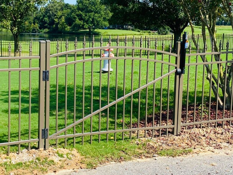 Metal park gate with diagonal brace, set in a grassy fenced area with trees in the background